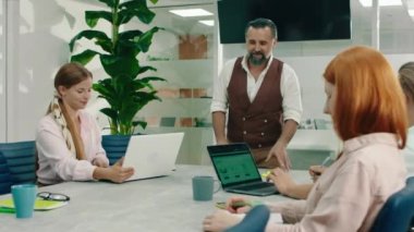 Kahverengi takım elbiseli yaşlı bir patron toplantı sırasında takım arkadaşlarına liderlik ediyor ve çok modern görünümlü bir ofiste iş problemlerini tartışıyor. 4k