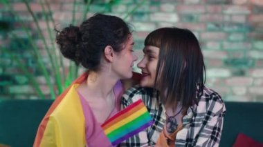 Oturma odasında kanepede kameraya yakın çok sevimli lezbiyen çift birbirlerine sarılıyorlar ve el ele tutuşuyorlar LGBT gökkuşağı bayrağı