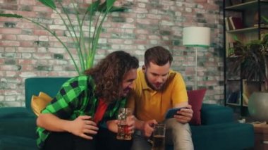 At home on the sofa handsome men watching together a sport match on the smartphone while holding some beers in hands then they getting excited for victory and cheers with the beers. 4k