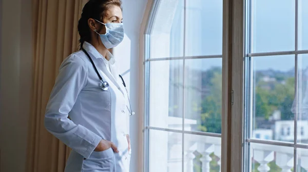 In the hospital mature doctor woman with a protective mask have a break time she came to the panoramic window and thinking.