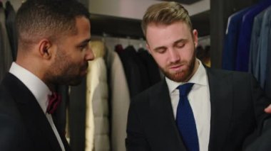 Yakışıklı damat düğün için en iyi ve en iyi takımı seçmeye çalışıyor. Koleksiyondaki yeni takıma bakan tezgahtar adamla birlikte. 4k