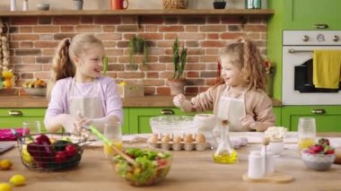 Very charismatic and beautiful two small sisters girls have a great time together at the kitchen island they preparing the dough for a delicious dessert together. 4k