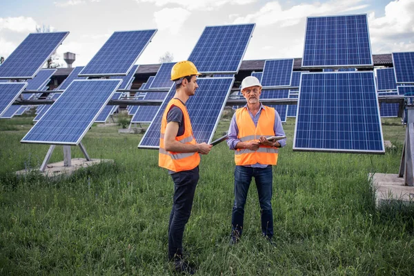 two young workers in overalls and helmets check the installed solar panels. Green electricity concept.
