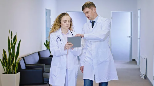 The main doctor man and his colleague female doctor walking hospital corridor and discussing the future treatment of the patient they stopping at reception to continue to analyzing the diagnostic