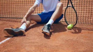 Outdoor on the clay court concept of professional sport tennis player guy sitting down beside the tennis net holding his racket and looking to the camera smiling large. Shot on ARRI Alexa Mini