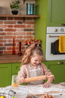 Happy and pretty little girl at the kitchen island using the roller and add some flour over the dough she happy take some juice and drink in front of the camera. Portrait