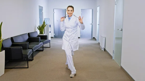 Charismatic lady doctor with a large smile dancing excited in the modern hospital corridor she looking straight to the camera.