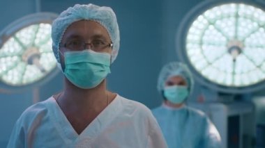 A handsome male surgeon with glasses is in a medical hospital room with a nurse standing behind him in very good lighting