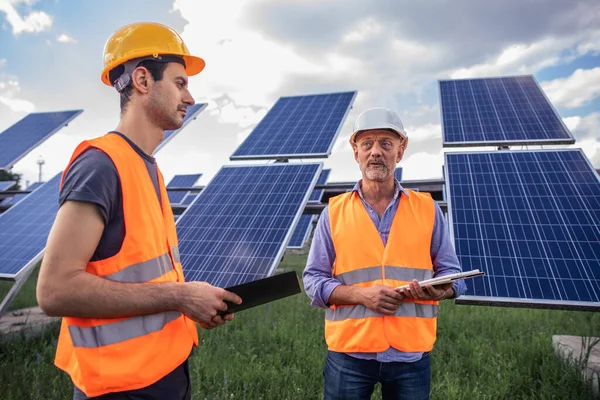 Medium shot of two electrical workers reviewing documents on a tablet during an inspection inbetween long rows of photovoltaic solar panels. Portrait