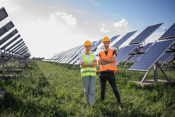 At large photovoltaic solar farm two ecological engineers young looking posing to the camera in a safety uniform they smiling large and crossing hands.