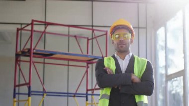 Attractive MAN architect posing in front of the camera crossing hands and looking straight at construction site he wearing helmets and protective glasses.