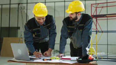 One engineer and constructor man at construction site analysing the plan of work working over the plan one of them take some notes wearing safety equipment and uniform. 4k