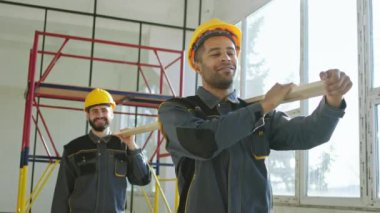 Modern construction site at industrial building in construction two changed multiracial constructors carrying a large wooden to the place of work. 4k