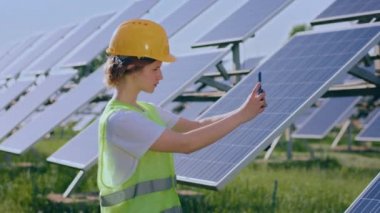Attractive woman ecological engineer with safety equipment at photovoltaic solar farm taking pictures of solar panels.