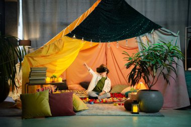 The boy chilling together inside a blanket fort while playing with a virtual reality headset and laughing.