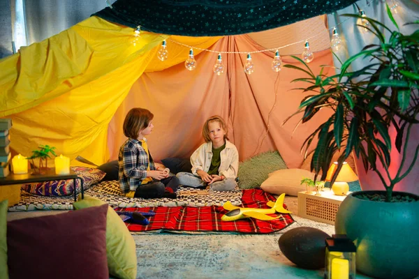 Two young boys, one brunette and one blonde are playing inside a tent that different colors.