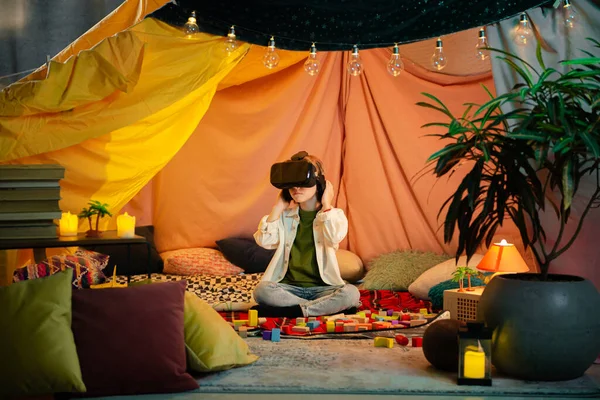 The boy are hanging out in a children s blanket tent indoors while playing with a vr headset and enjoying their time laughing together.