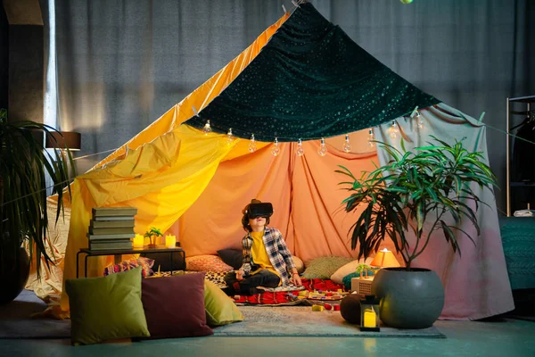 The boy are inside a massive and brightly colored indoor blanket fort while playing together with a vr headset.