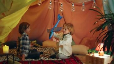 Two cute boys are playing with airplanes inside a blanket tent where there are lightbulbs hung across the top and it looks very comfy. 4k
