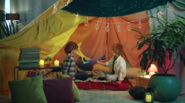 Two junior boys are playfully playing with yellow and blue airplanes inside a blanket fort which has very colorful bright blankets.
