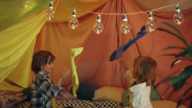 Two young guys with longer hair are having fun together in a comfy indoor tent, there are pillows behind them and blankets everywhere.