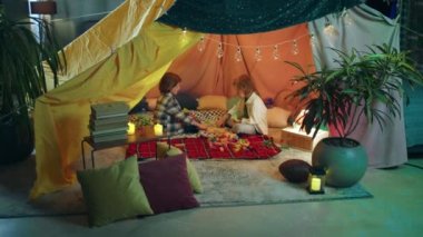 Inside a very hippy looking sheet tent with plants there are two boys curiously playing together with different colored block.