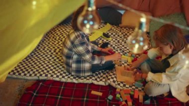 A zooming in shot of two boys focusing on building a tower together with blocks while sitting down in a luminous indoor sheet fort.