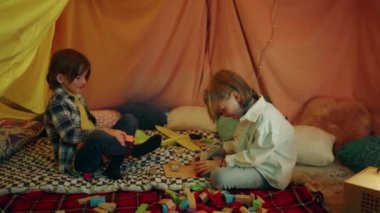 Two younger boys are smiling and laughing with eachother while throwing blocks at one another and sitting inside a huge tepee made out of sheets.