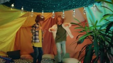 Two delightful looking boys made a blanket tent indoors and now they re both jumping and laughing together inside it in a very happy atmosphere.