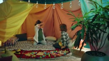 Two boys are chilling together inside a blanket fort while playing with a virtual reality headset and laughing.