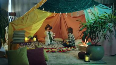 Two little boys are inside a massive and brightly colored indoor blanket fort while playing together with a vr headset.