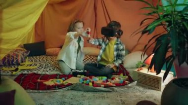 One little blond boy is showing things to another brunette boy who is wearing a vr headset inside a comfortable sheet fort.
