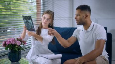 In a modern clinic at doctors office the patient guy sitting on the sofa together with the doctor they discussing about the x-ray doctor holding the result of x-ray on hands. 4k