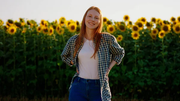 A young foxy looking woman is wearing a checkered blouse and standing in front of a sunflower field showing her thumbs up while smiling widely at the camera. Portrait