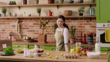 Excited dancing lady at home while preparing the dinner she smiling large at the kitchen island and wear the kitchen apron.