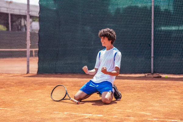 Professional tennis match young guy hitting the ball the sitting on the knees and getting very excited after he win the match outdoor on the clay court. Portrait