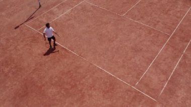 Afro American guy professional tennis player playing tennis in a sunny day on the large tennis court outdoor taking video with drone birds eye view.