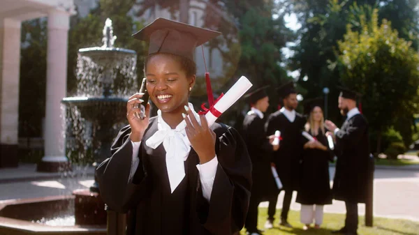 卒業式の日にとても美しい黒の女性は 電話を取り 彼女はすべてのグループで議論する他の学生を背景に卒業した後 彼女が話すと同時に笑顔誰かを呼び出します — ストック写真