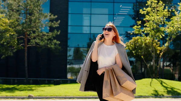 Beautiful ginger hair lady with some shopping eco bags walking in front of the camera and speaking on her smartphone with someone she wearing sunglasses.