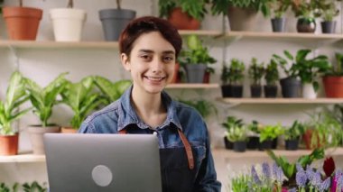 Çiçekçi dükkanındaki güzel bayan çiçekçi elinde dizüstü bilgisayarla kameranın önünde poz veriyor. Gülümsüyor ve dik duruyor. ARRI Alexa Mini 'de çekilen