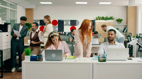 A beautiful ginger haired manager is checking up on her assistants work on their laptop and giving them advice while in the back a group of three workers are talking about work. Portrait