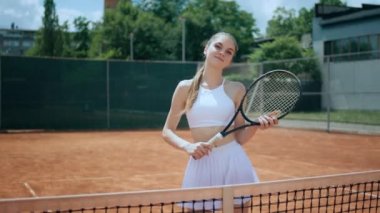Güzel profesyonel tenisçi kadın kameranın önünde elinde tenis raketiyle poz vermekten ve tenis kortunun önünde gülümsemekten mutlu. ARRI 'de çekilen