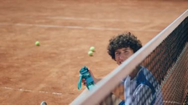 Açık hava kil sahası kameranın önünde profesyonel tenis oyuncusu genç tenisçinin yanında kameraya bakıp gülümsüyor ve elinde bir şişe su tutuyor. ARRI 'de çekilen