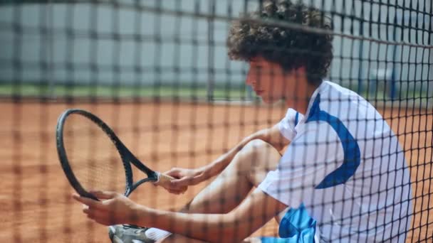 カリスマ的な若いテニス選手のビデオを撮影し 彼はテニスネットの横にある粘土のコートに座って カメラを探してラケットで遊んでいました Arriで撃たれた — ストック動画