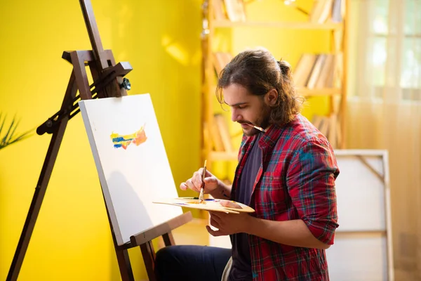 A beautiful man with a beard and a manbun is concentrating on the art he is painting with lots of colours on his canvas.