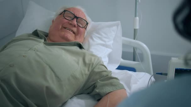 病院のベッドで寝そべっている間に笑顔の先輩は 彼の隣に座っている彼の娘を回復し 彼をサポートし 奨励しています — ストック動画