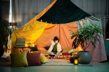 The boy chilling together inside a blanket fort while playing with a virtual reality headset and laughing.