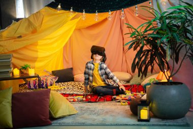 One little blond boy is showing things to another brunette boy who is wearing a vr headset inside a comfortable sheet fort.