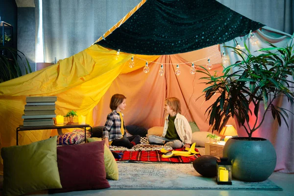 Two young cute boys are playing inside a nice comfy tent that has a hippie vibe to it, the tent is an indoor one.