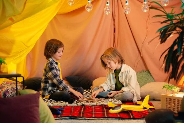 Two young boys which are similar in age are playing inside an indoor tent and messing around with a flashlight.